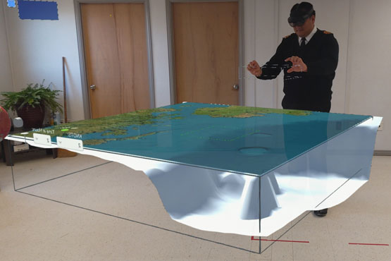 A Royal Canadian Navy member wearing an augmented reality headset gestures with his hands at a virtual three-dimensional model, showing the ocean and coastline.