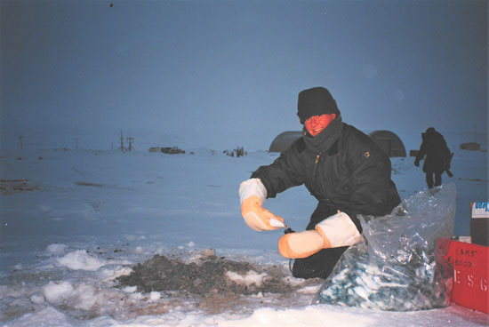 A Canadian Armed Forces member wearing thick mittens and a toque collects a soil sample in a snow-covered field.
