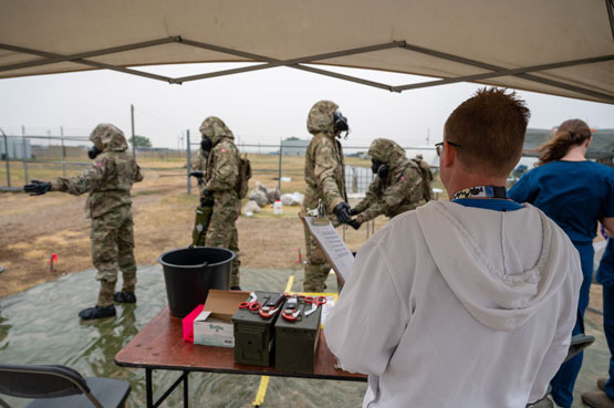 Soldiers in protective gear lined up in front of a table being observed by a man with a clipboard