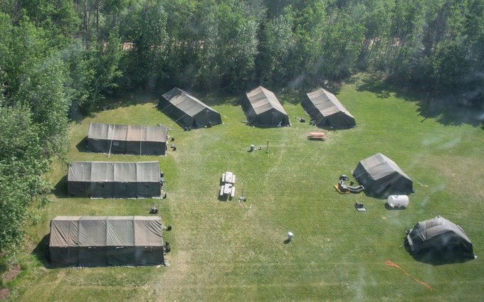 Green military tents are arranged in a semi-circle in a field, surrounded by forest.